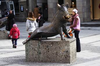 A Person Interacts With A Bronze Boar Statue In A Bustling Munich City Square, Where Passersby, Including A Child In A Red Jacket, Go About Their Day.