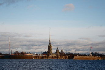 A Golden Spire Pierces The Sky Above A Historic Fortress On A Waterfront In St. Petersburg, Under A Vast Expanse Of Clouds Tinged By The Soft Light Of Either Dawn Or Dusk.