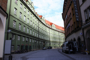 A Narrow Munich Alleyway With Cobblestone Streets Flanked By Green And Beige Traditional Buildings, A Parked Van On The Right, And Bicycles Lined Against The Wall Under Clear Skies.