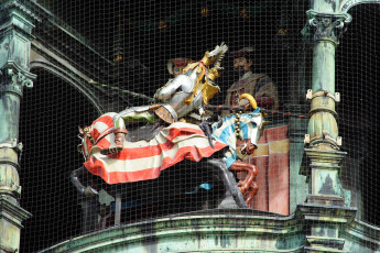 A Vibrant, Medieval Knight Statue With A Golden Lance On Horseback, Accompanied By Figures In Period Attire, All Ensconced Behind Protective Netting Atop An Ornate, Green-Roofed Structure In