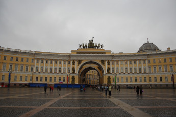 The General Staff Building Arch Glows Warmly In Yellow Hues, While A Horse-Drawn Chariot Perches Atop, Overlooking A Cloudy Sky And A Scattering Of Visitors In St. Petersburg'S Palace Square.