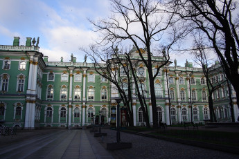 A Baroque-Style Green And White Palace In St. Petersburg, With Ornate White Sculptures And Windows Under A Blue Sky, Flanked By Bare Trees, In A Cobblestone-Paved Courtyard.