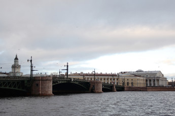Overcast Sky Over A Wide River With A Classic Multi-Arched Bridge Leading To Historic European-Style Buildings And A Prominent Tower In The Background, Capturing A Serene Yet Somber Urban Landscape In St.