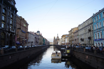 A Tranquil Evening Settles Over The St. Petersburg Canal Lined With Historic Buildings, Where A Single Tour Boat Floats Gently, Under The Gaze Of A Distant Ornate Cathedral.