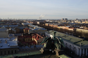 A Panoramic View Of St. Petersburg With Historic Architecture, Featuring Rooftops Under A Clear Sky, With Industrial Smokestacks In The Distance, As Seen From An Elevated Position.