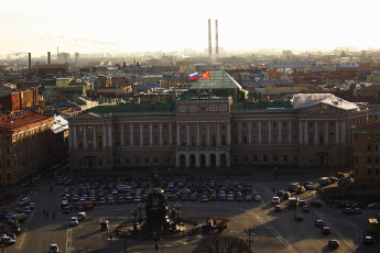 Aerial View Of A Historical Square In St. Petersburg At Sunset, Featuring A Grand Building With A Green Rooftop And Flag, A Prominent Statue, And Neatly Parked Cars, Against A Cityscape Backdrop With