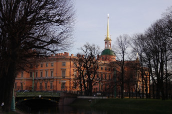 Historic Orange Building With A Green Dome And Golden Spire Stands By A Serene Water Canal In St. Petersburg, With A Bridge In The Foreground Amidst Leafless Trees Under A Soft Dusk Sky.