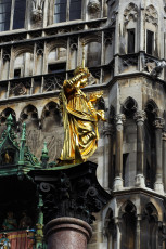 A Gilded Statue Gleaming In The Sunlight, Possibly A Historical Or Religious Figure, Stands Before An Ornate Gothic Building With Detailed Stone Facades And Architectural Decorations In Munich.