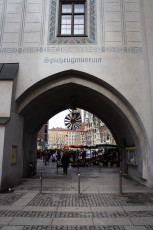 An Arched Entrance With The Sign &Quot;Spielzeugmuseum&Quot; Leads To A Vibrant City Square In Munich, Where A Colorful Market Is Visible And A Historic Building With Intricate Façades Stands In The Background