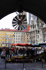 An Archway Frames A Bustling Munich City Square Where Vendors Sell Vibrant Flowers Beneath A Historical Clock, With Gothic Architecture And Urban Facades Blending In The Backdrop.
