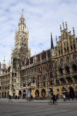 The Gothic Revival Architecture Of The New Town Hall Graces Munich'S Marienplatz, Under A Clear Blue Sky, With Pedestrians And Cyclists Enjoying The Spacious Square.