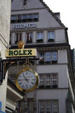 A Golden Rolex Sign With An Ornate Clock Extends Over The Sidewalk In Front Of A Multi-Story Building With The Name &Quot;Hirmer&Quot; On Its Facade, Suggesting A Luxury Watch Retailer In An Urban