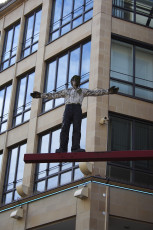 A Life-Size Sculpture Of A Human Figure With Arms Outstretched Stands On A Red Beam, Mounted On The Facade Of A Modern Building In Munich With Large Windows And Stone Cladding, Creating An Eye