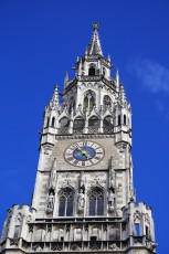A Clear Blue Sky Provides A Backdrop For The Ornate, Gothic Tower Of A Historic Building In Munich, Featuring Intricate Spires, A Large Clock Face, And Statues Nestled In Its Architecture.