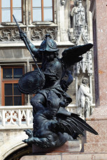 A Bronze Statue Of A Warrior Angel, Wielding A Sword And Shield, Stands Defiantly With Wings Outstretched Against A Backdrop Of Ornate Gothic Architecture In Munich.