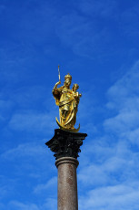 A Golden Statue Of A Robed Female Figure Holding A Spear And Shield, Standing Atop A Tall Column Against A Clear Blue Sky In Munich.