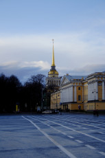 A Quiet, Expansive City Square In St. Petersburg With Crisscrossing White Lines On The Asphalt, Leading To A Historic Building With A Golden Spire Piercing A Blue Sky Scattered With Clouds.