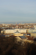 A Panoramic View Of St. Petersburg'S Cityscape With Historic Buildings Under A Soft Sky, Industrial Cranes In The Distance, And The Foreground Featuring Autumnal Trees, Suggesting A Blend Of Urban Life And