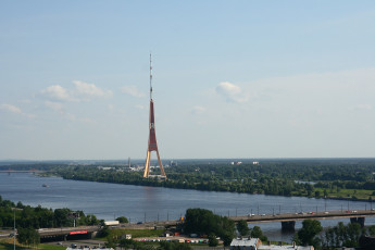 A Bridge Over A River With A Tall Tower In Riga From Above.